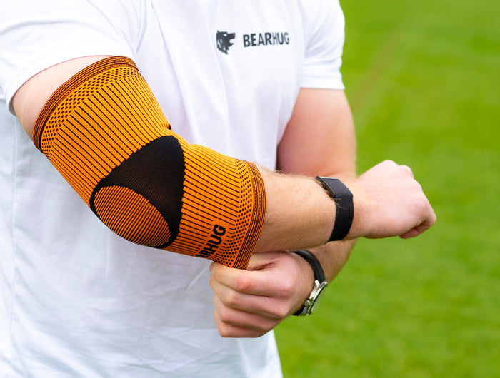 Person wearing a white shirt and black shorts with an orange elbow support on a grassy field.